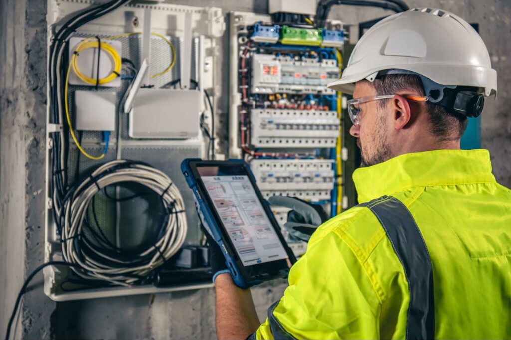 An electrical technician operating a switchboard with fuses, utilizing a tablet while employing data security measures.