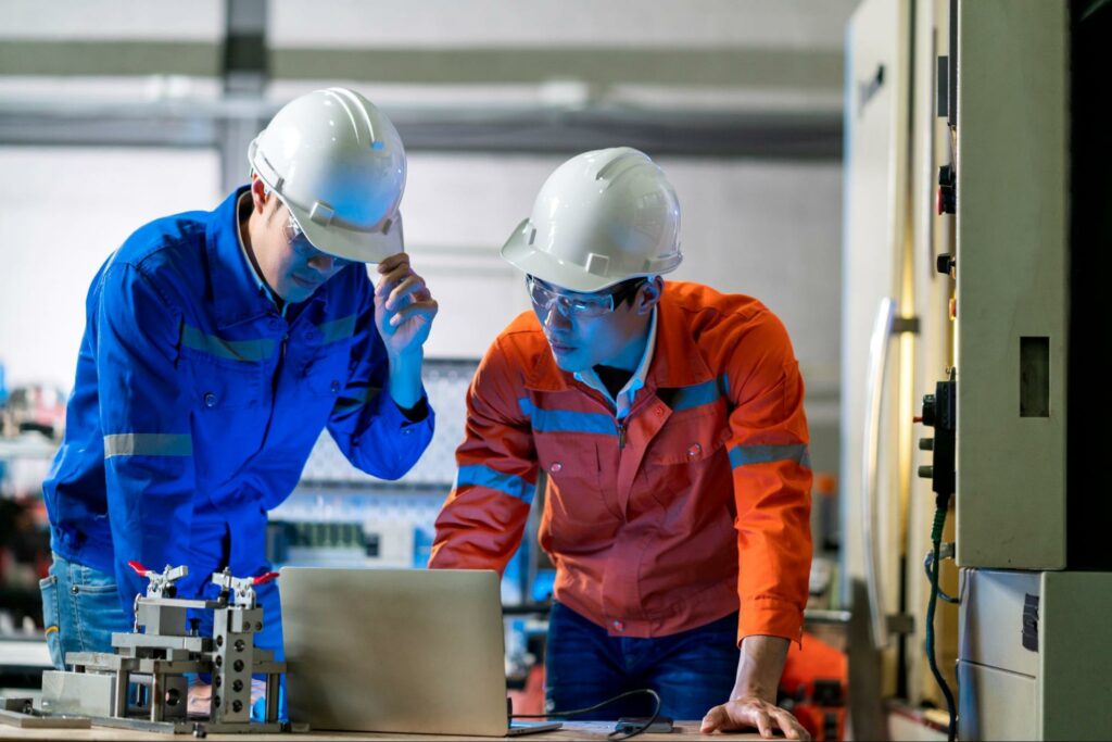 Two cybersecurity engineers discussing and brainstorming beside a CNC machine in the factory, with one coworker explaining solutions.