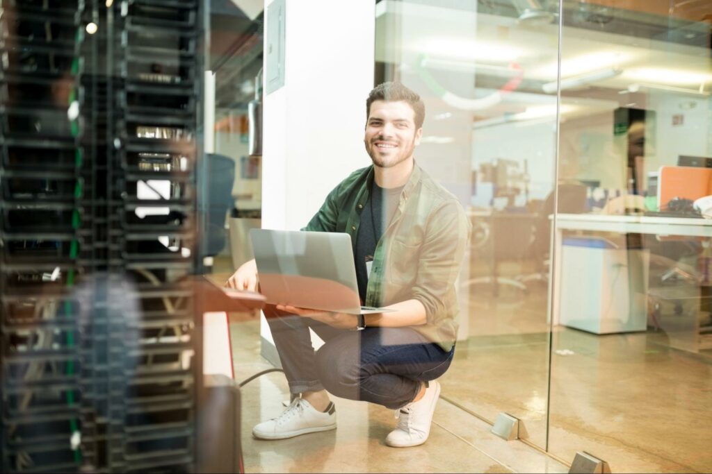 Smiling male network engineer with laptop in a datacenter, merged with hardware-based cybersecurity for a harmonious blend of expertise and digital protection.